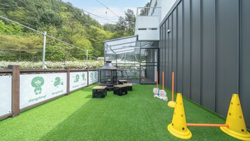 Un espacio al aire libre con césped artificial, un recinto de cristal y un parque infantil con conos amarillos.