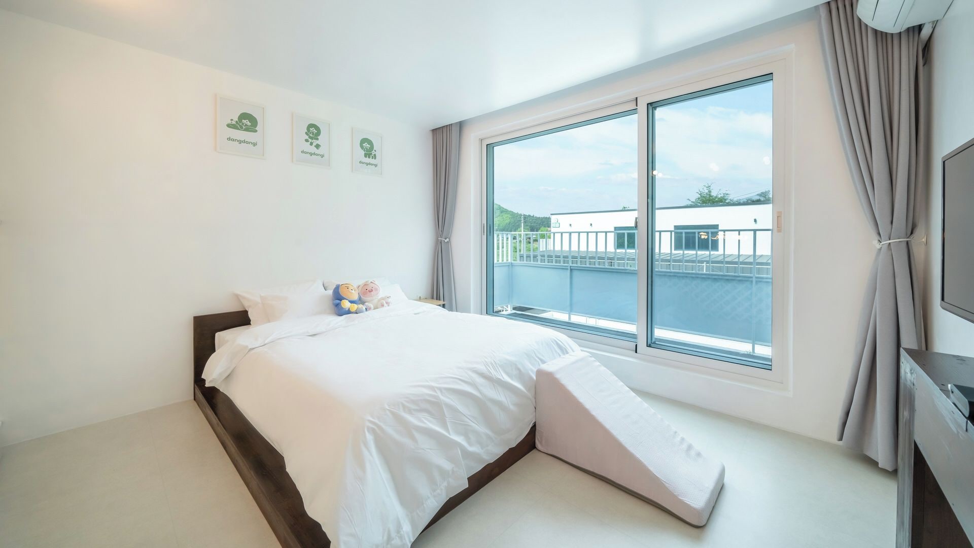 Habitación de hotel con cama, televisor y ventana con vista a un edificio y vegetación.