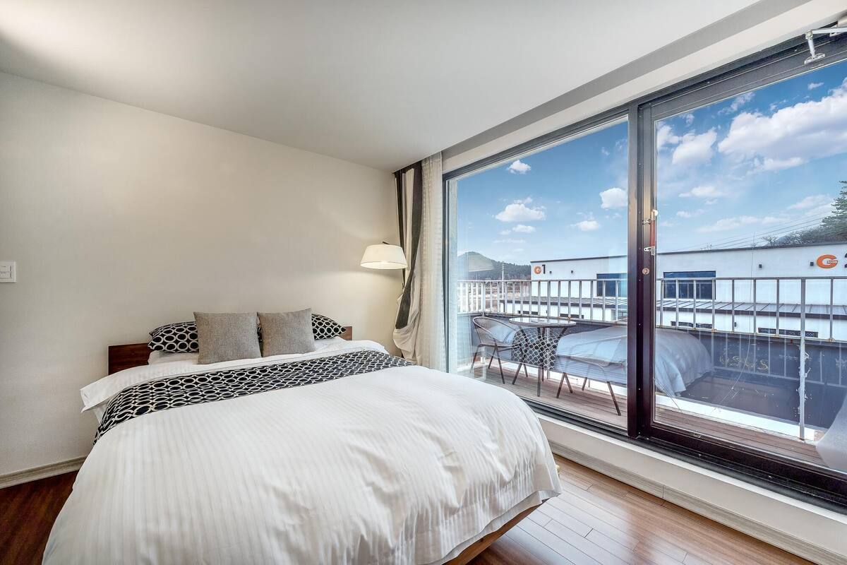 Une chambre d’hôtel avec une grande fenêtre, un lit recouvert d’une literie à motifs, une table de chevet avec une lampe, et une vue sur un bâtiment situé de l’autre côté de l’eau.