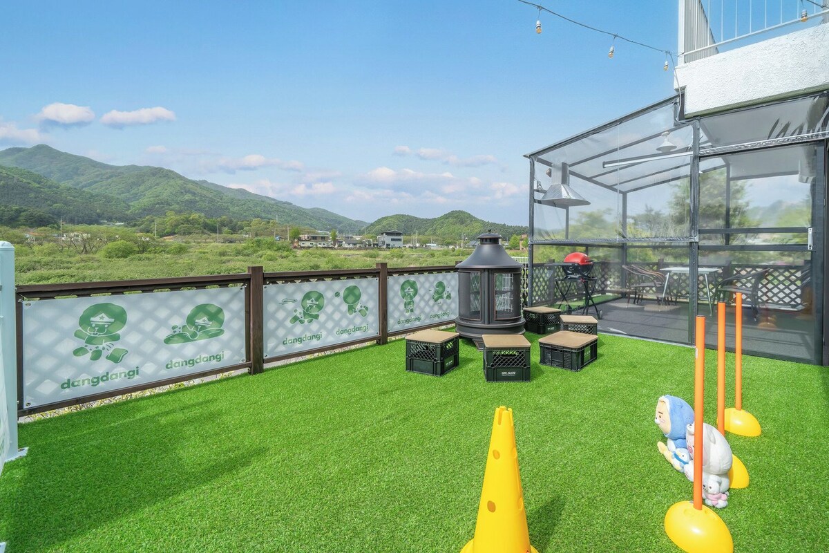 A rooftop terrace with artificial grass, outdoor furniture, a grill, and a view of the mountains.