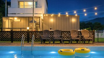 Zona de piscina con sillas, flotadores hinchables, una terraza de madera y un edificio con luces colgantes.