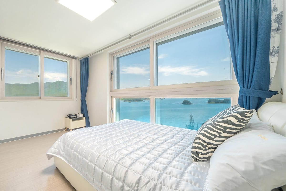 Un dormitorio con una cama grande, dos ventanas, una mesita de noche y vista al mar.