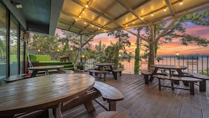 Un patio al aire libre con mesas y bancos de madera, una zona de césped verde y vistas a un atardecer sobre el agua.