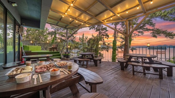 Un espace extérieur aménagé pour les repas, avec des tables et des bancs en bois, un barbecue et une vue sur le coucher de soleil au-dessus d’une étendue d’eau.
