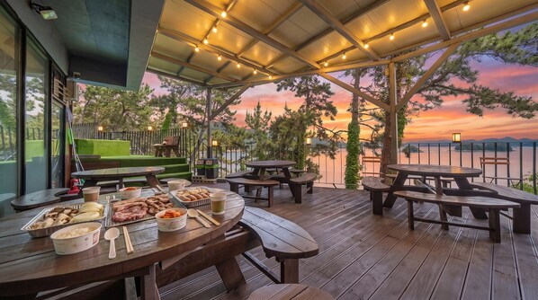 Un espace extérieur aménagé pour les repas, avec des tables et des bancs en bois, un barbecue et une vue sur le coucher de soleil au-dessus d’une étendue d’eau.