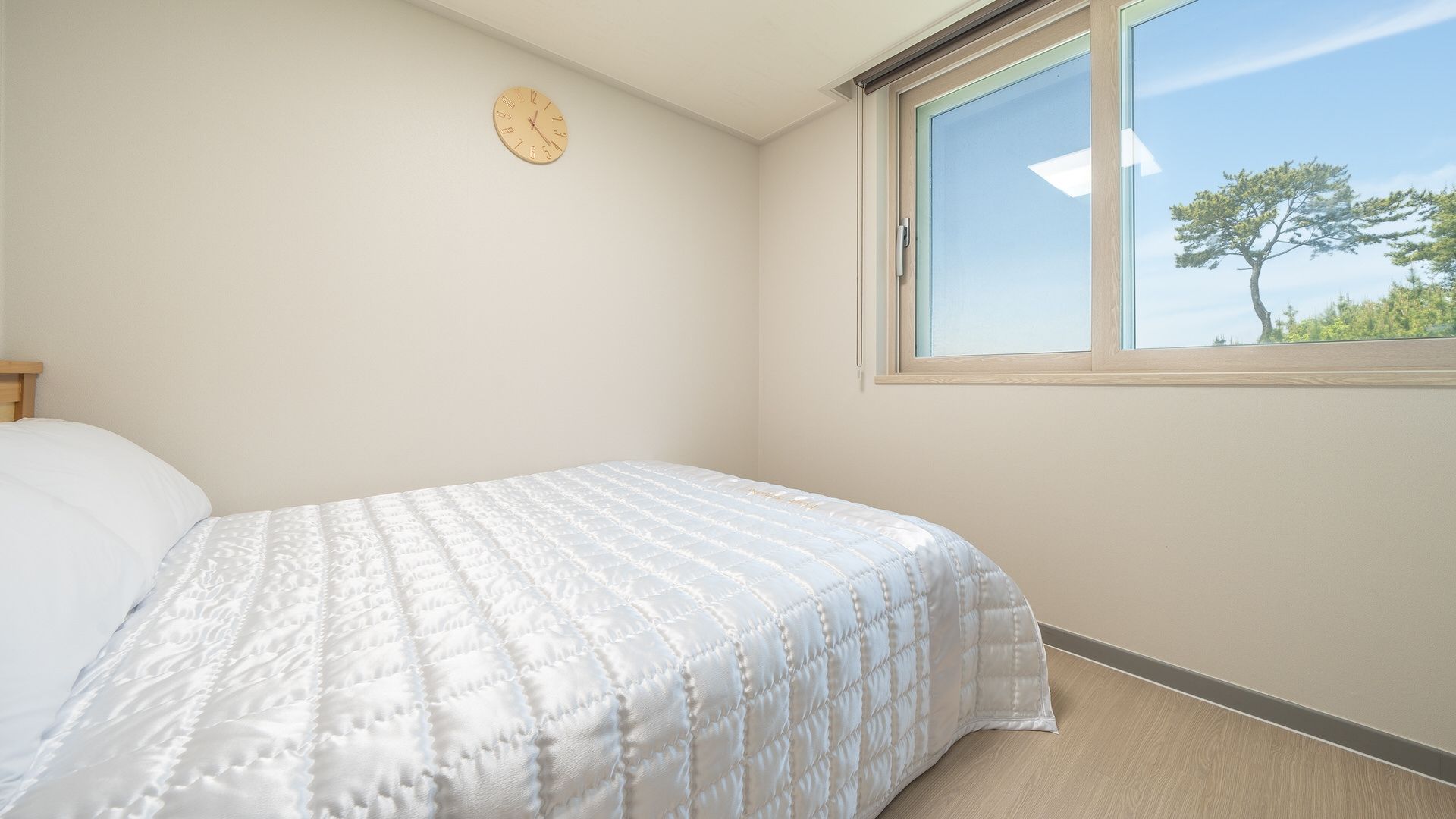A bedroom with a bed, a wall clock, and a window with a view of trees.