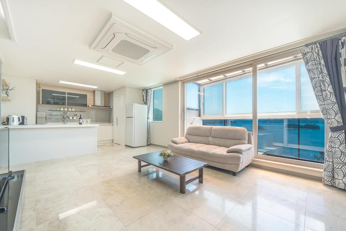 Una sala moderna con sofá y mesa de centro, una cocina con armarios blancos, y un ventanal con vistas al mar.