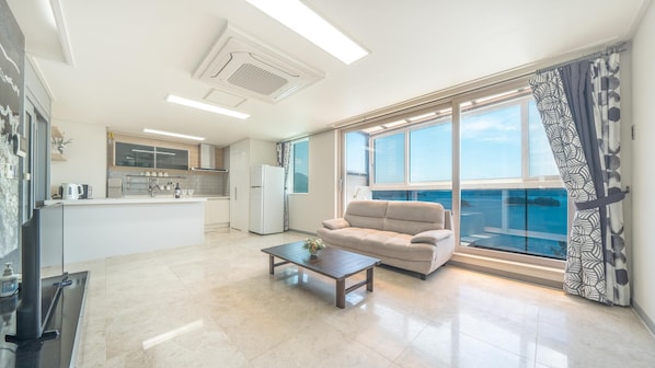 Una sala moderna con sofá y mesa de centro, una cocina con armarios blancos, y un ventanal con vistas al mar.