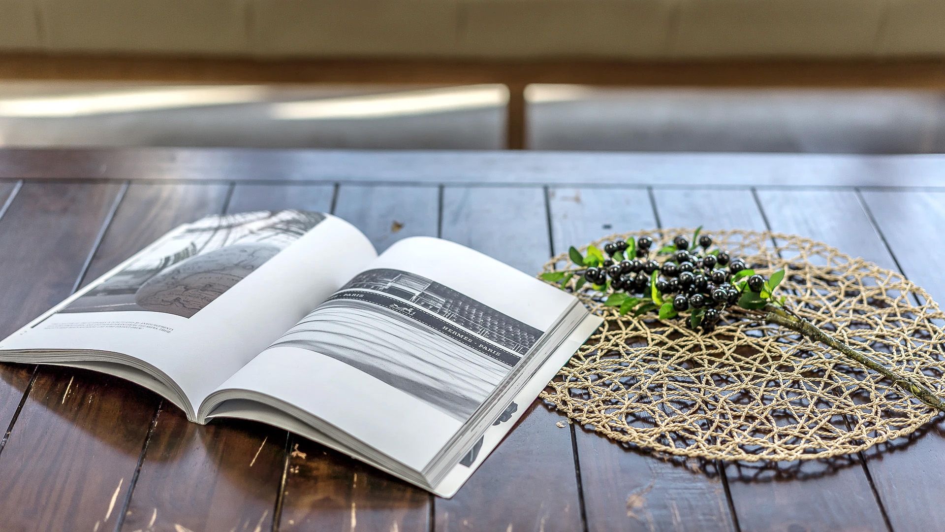 An open book on a wooden table with a woven placemat and a small branch with berries.