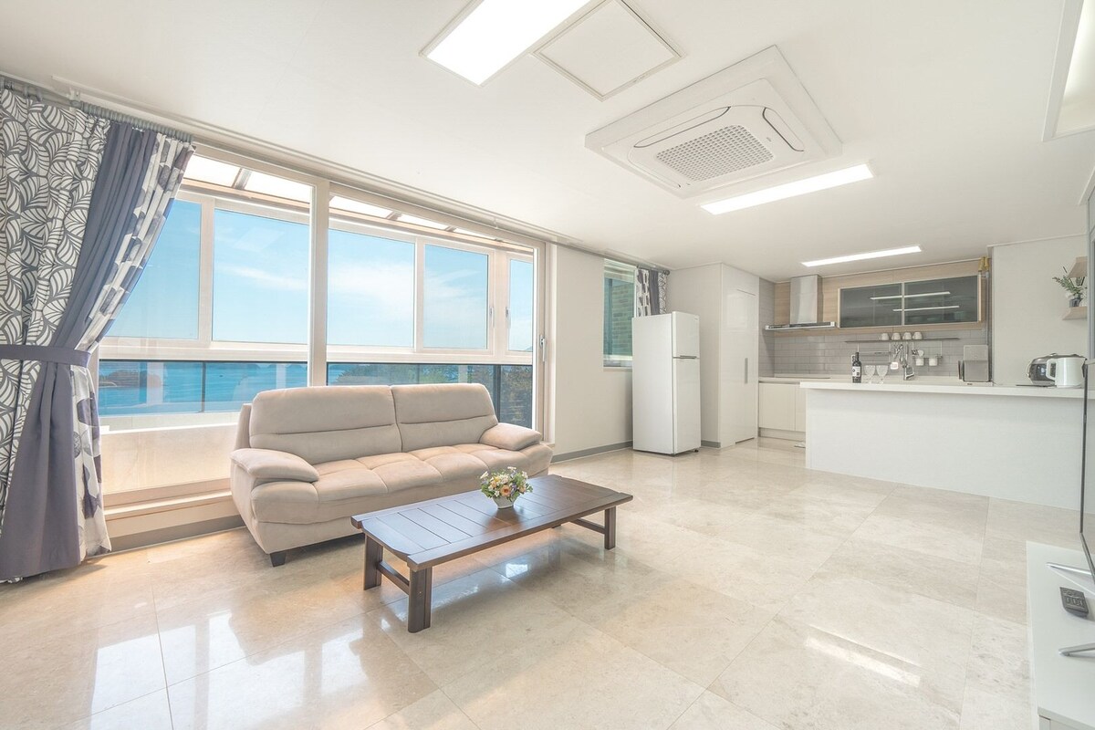 Un salon moderne avec un canapé, une table basse et une vue sur la mer grâce à de grandes fenêtres.