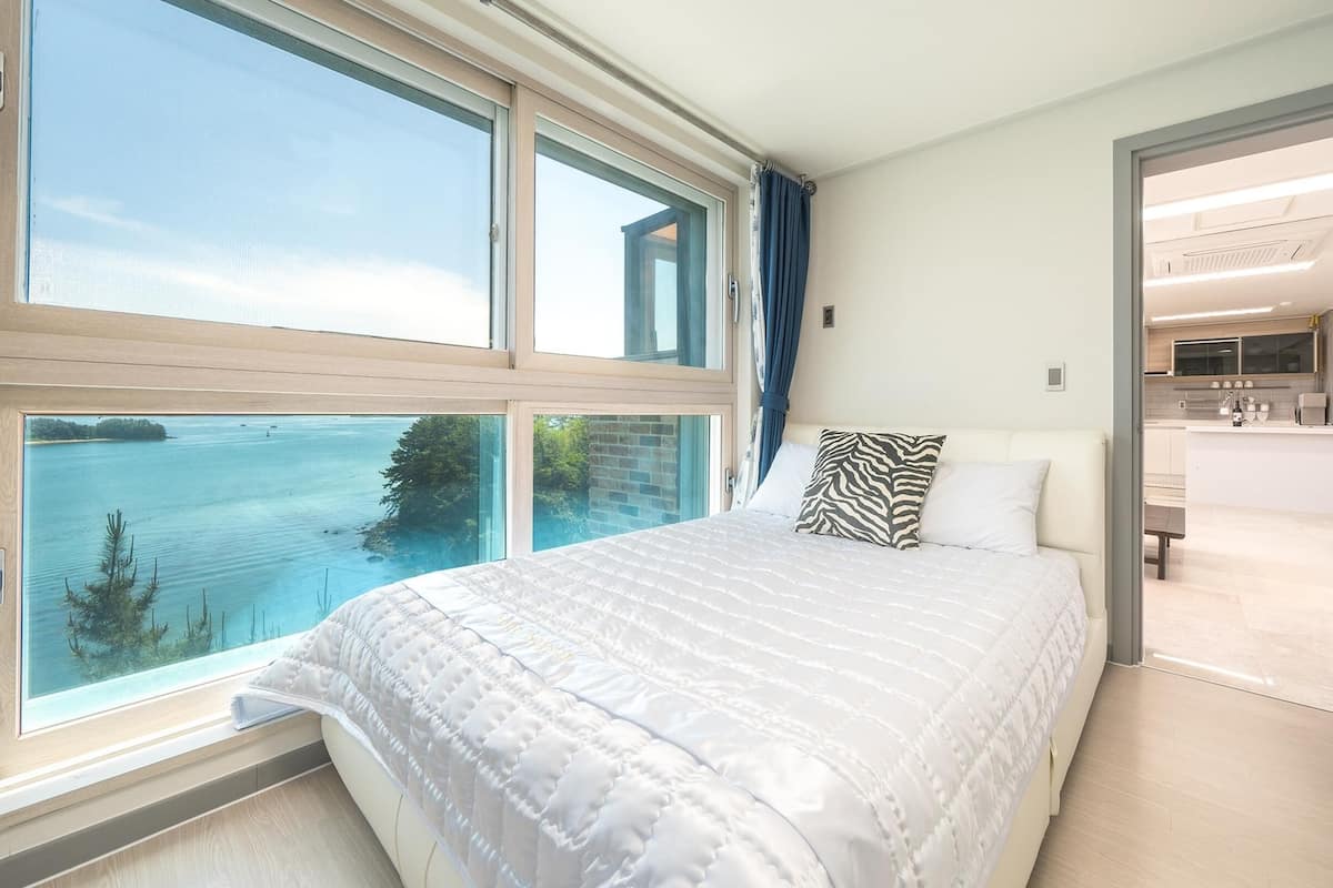 Un dormitorio con un ventanal que da a una masa de agua, una cama con ropa de cama blanca y vistas a una cocina moderna.
