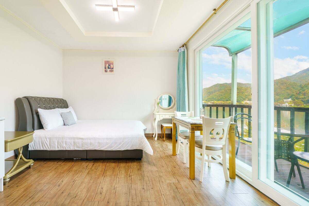 Une chambre avec un lit, un bureau et une chaise, une fenêtre donnant sur des collines et un balcon.