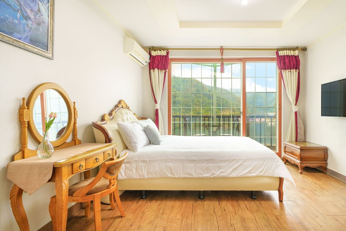 Une chambre avec un grand lit, une commode en bois, une coiffeuse avec un miroir et un téléviseur fixé au mur.