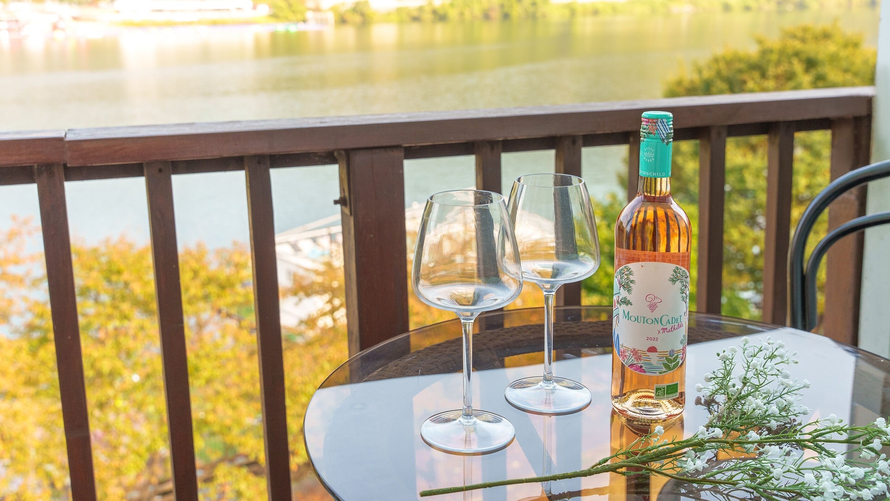 A table with two empty wine glasses and a bottle of wine on a balcony overlooking a body of water.