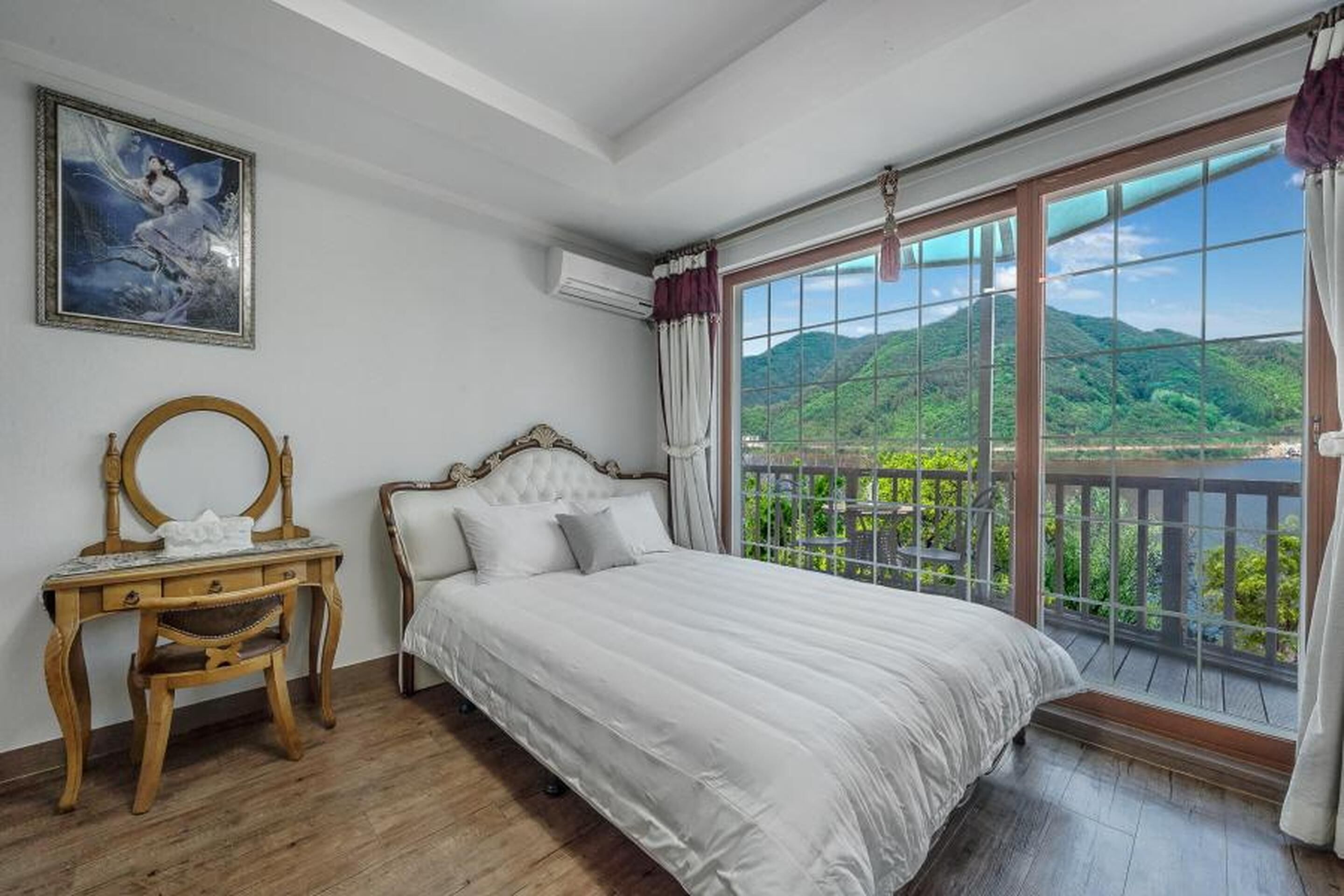 Een slaapkamer met een groot bed, een houten bureau, een stoel en uitzicht op een groen landschap door een groot raam.