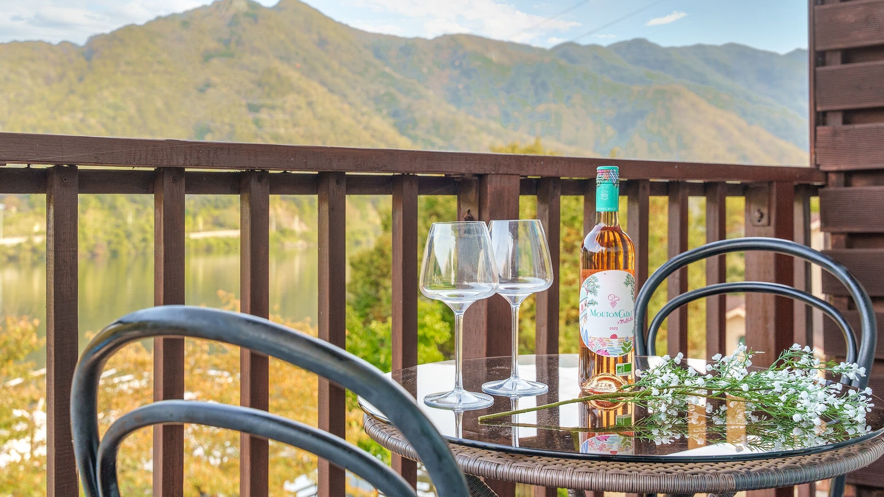 A glass table with two wine glasses, a bottle of Mouton Cadre, and a bouquet of flowers on a balcony overlooking a lake and mountains.