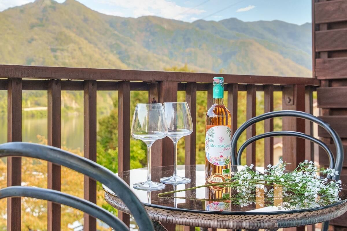 Une table en verre sur laquelle reposent deux verres à vin, une bouteille de Mouton Cadre et un bouquet de fleurs. La table est placée sur un balcon qui offre une vue sur un lac et des montagnes.