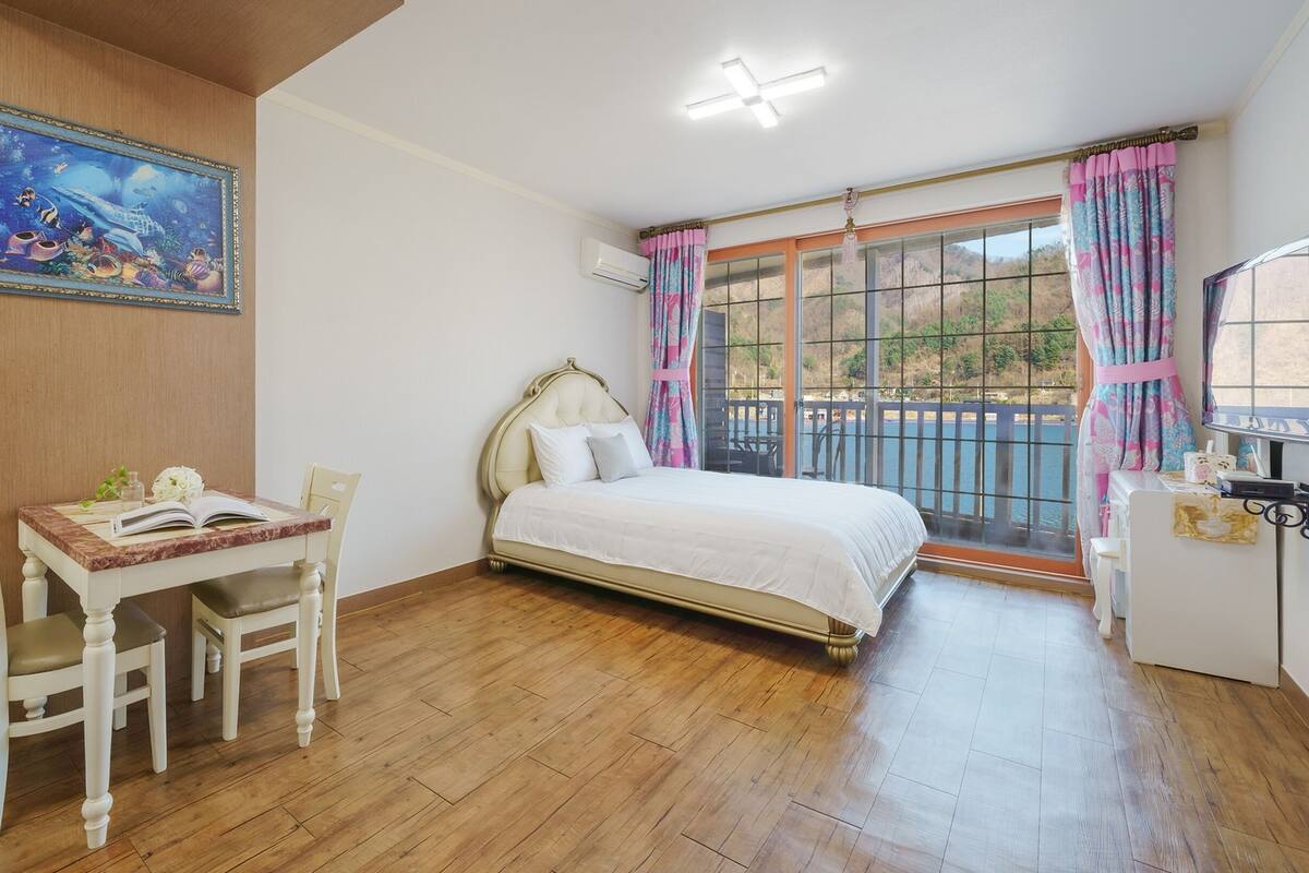 Une chambre à coucher avec un lit, un bureau avec des chaises, une fenêtre avec des rideaux et un tableau au mur.