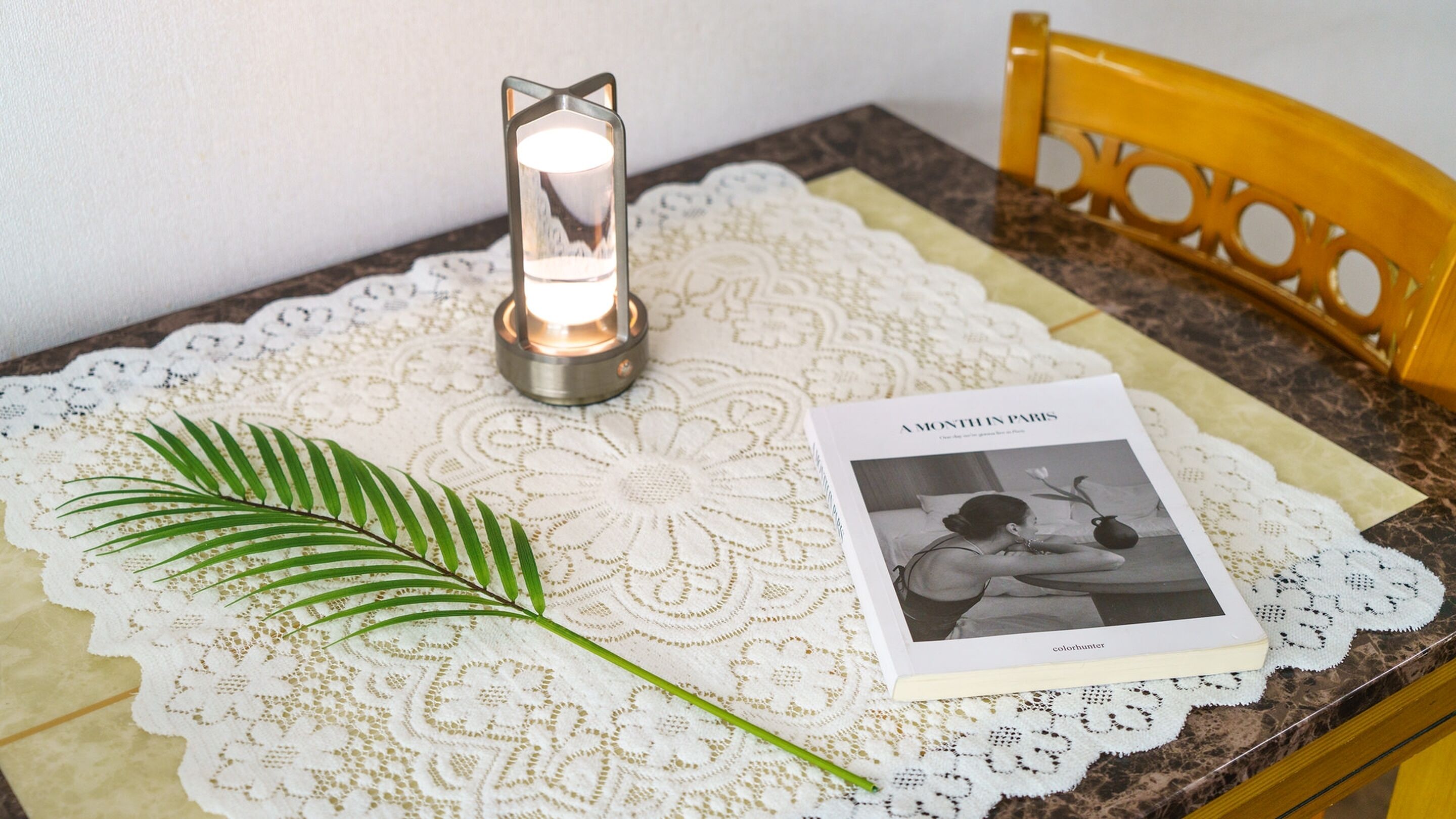 A table with a lace tablecloth, a candle, a book titled 'A Month in Paris', and a green leaf.