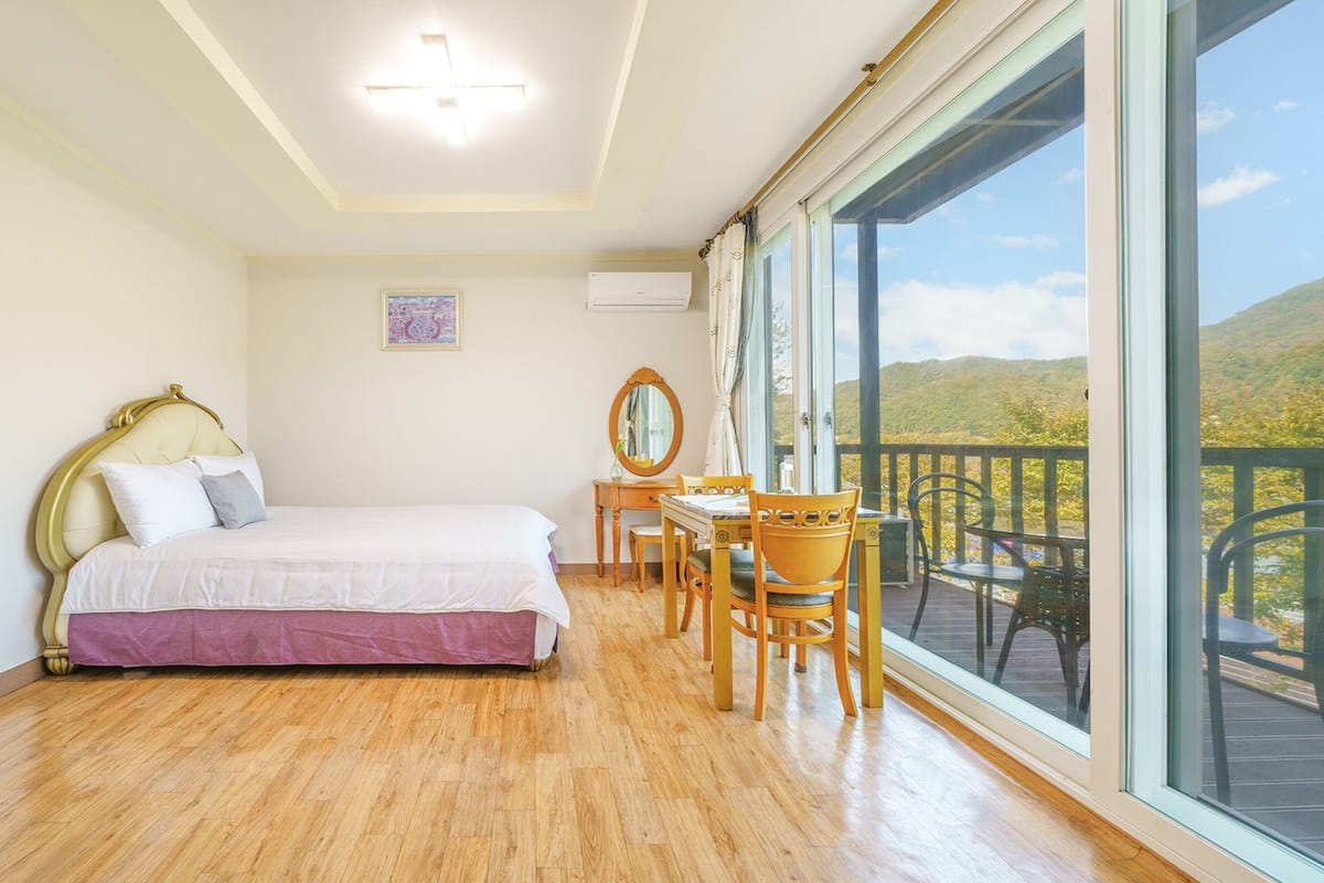 Une chambre avec un lit, un bureau et une chaise, une grande fenêtre donnant sur des collines, et un climatiseur fixé au mur.