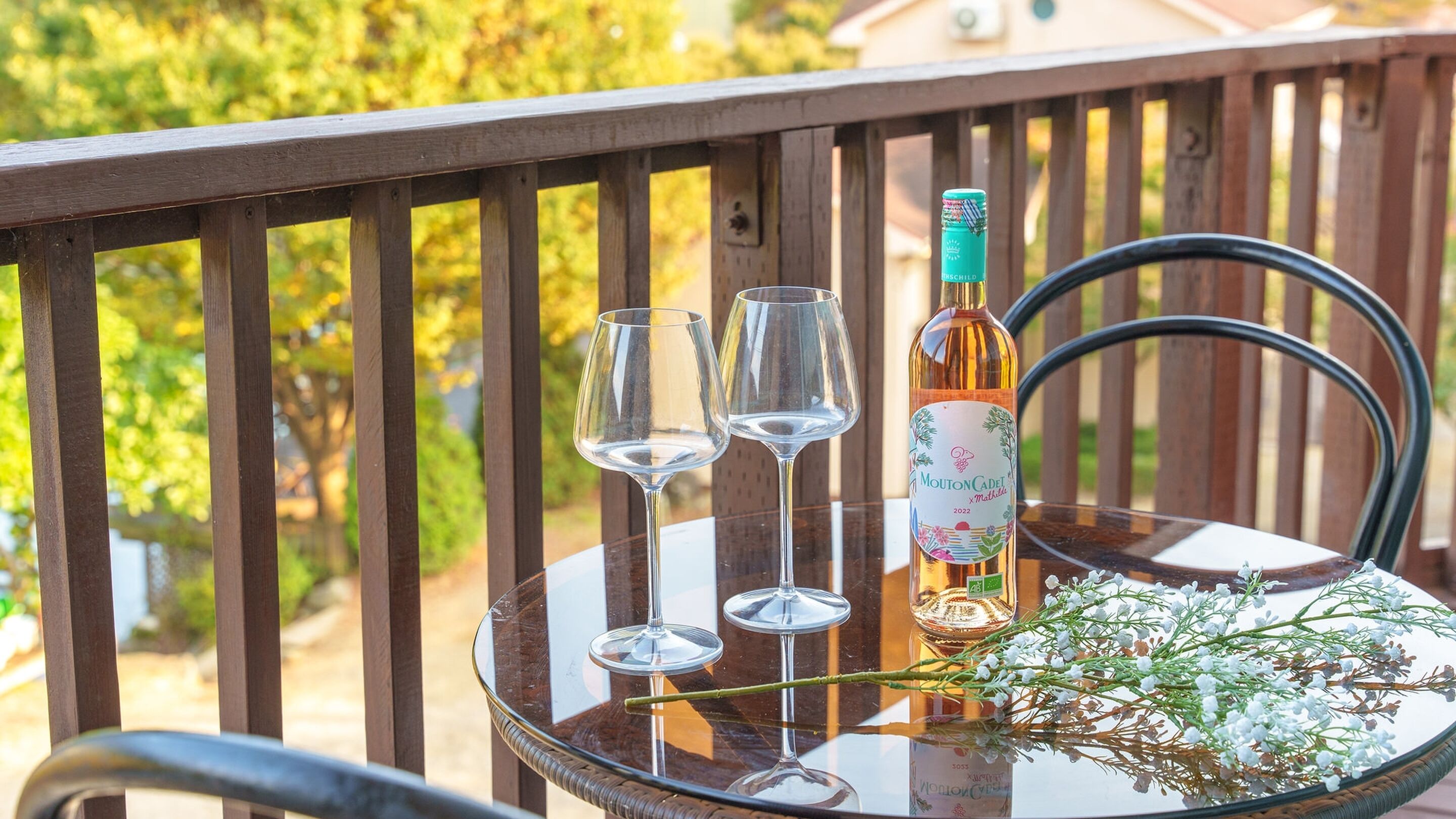 Una mesa de cristal con una botella de vino Mouton Cadet, dos copas de vino vacías y una rama con flores blancas en un balcón.
