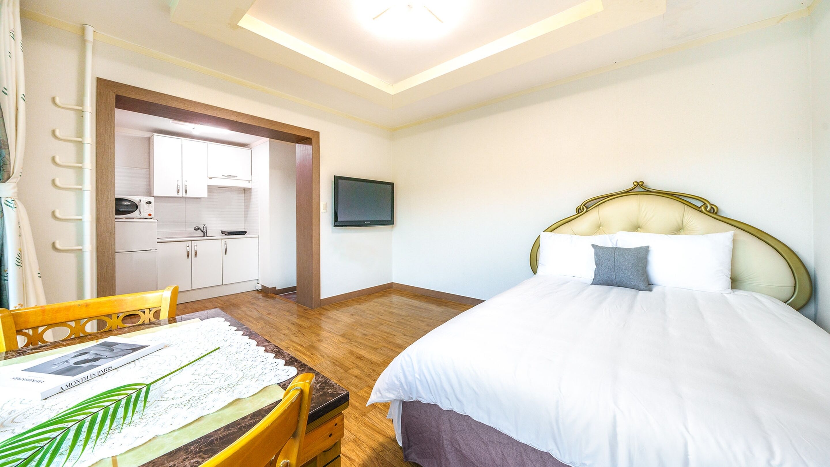A bedroom with a bed, a desk with a chair, a television on the wall, and a kitchenette in the background.