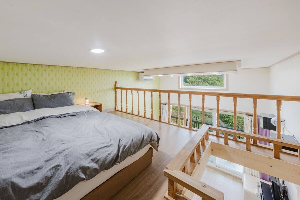 A bedroom with a bed, a nightstand, a wooden railing, and a window overlooking greenery.