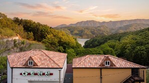 Un bâtiment doté d’un toit recouvert de tuiles et d’une enseigne avec des caractères coréens, entouré d’une végétation luxuriante et avec des montagnes en arrière-plan.