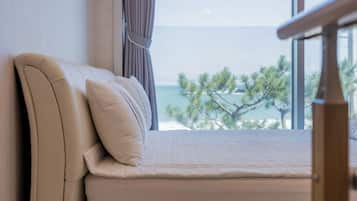 A bed with a headboard and pillows, positioned next to a window with a view of the sea and trees.
