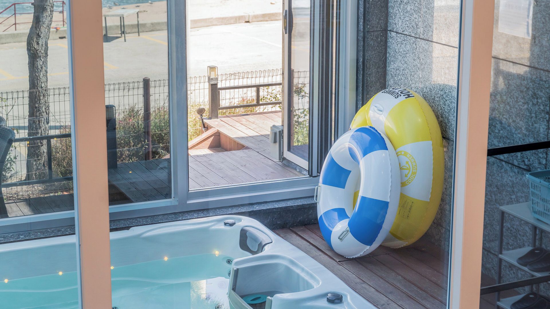 Una bañera de hidromasaje con cubierta de vidrio, dos flotadores hinchables, una terraza de madera y vista a la playa.
