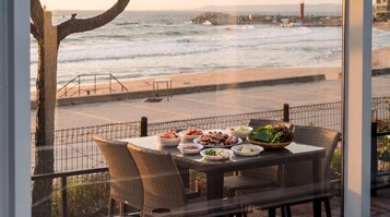 A dining table with various dishes and a wicker chair, overlooking a beach with the ocean and a lighthouse in the distance.