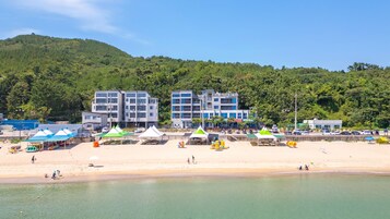 Una playa con una hilera de cabañas de playa, un edificio con el letrero "WOLF WOLF" y una colina boscosa al fondo.