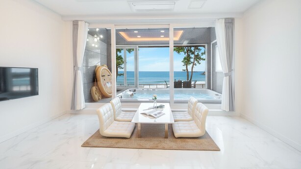 Un salon moderne doté d’une grande fenêtre donnant sur une plage, d’un coin salon avec une table et d’un téléviseur à écran plat fixé au mur.