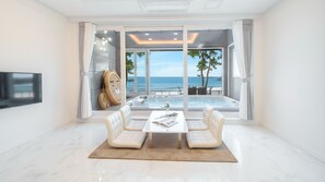 A modern living room with a large window overlooking a beach, a seating area with a table, and a flat-screen TV mounted on the wall.