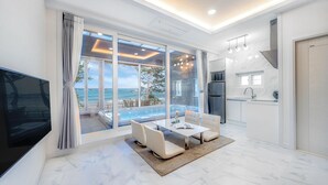 Un salon moderne doté d’une grande porte-fenêtre coulissante donnant accès à une zone piscine, d’une cuisine avec réfrigérateur et d’un téléviseur à écran plat.