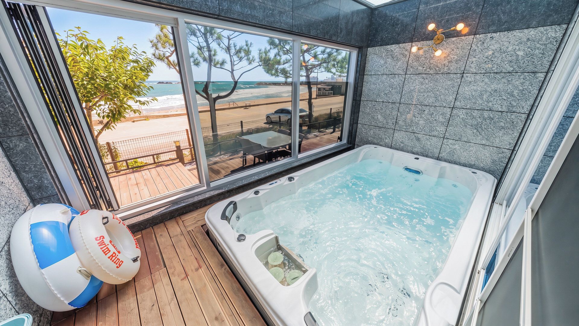 A modern bathroom with a large bathtub, a view of the beach, and a wooden deck outside.