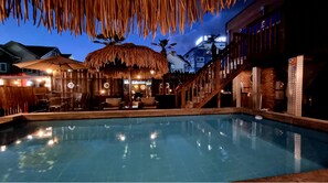 A pool area with a thatched-roof structure, lounge chairs, and a staircase leading to an upper level.