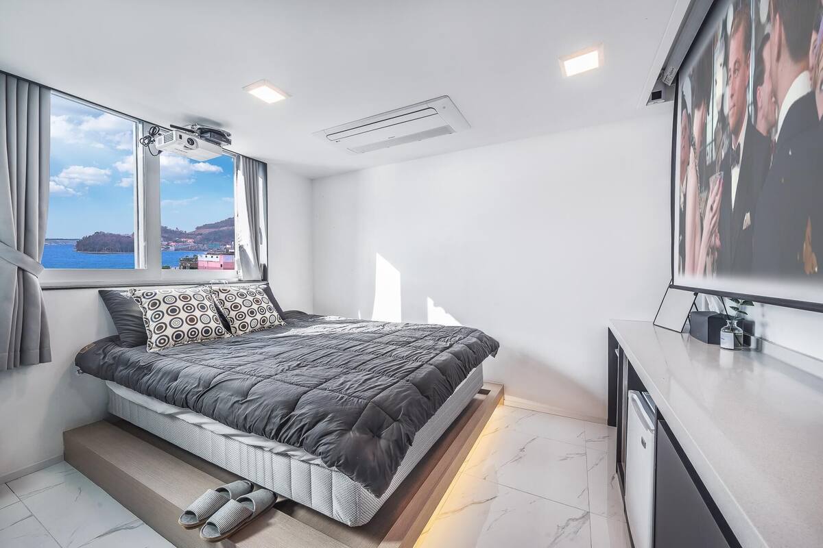 A modern bedroom with a bed, a TV, and a window with a view of a body of water and buildings.
