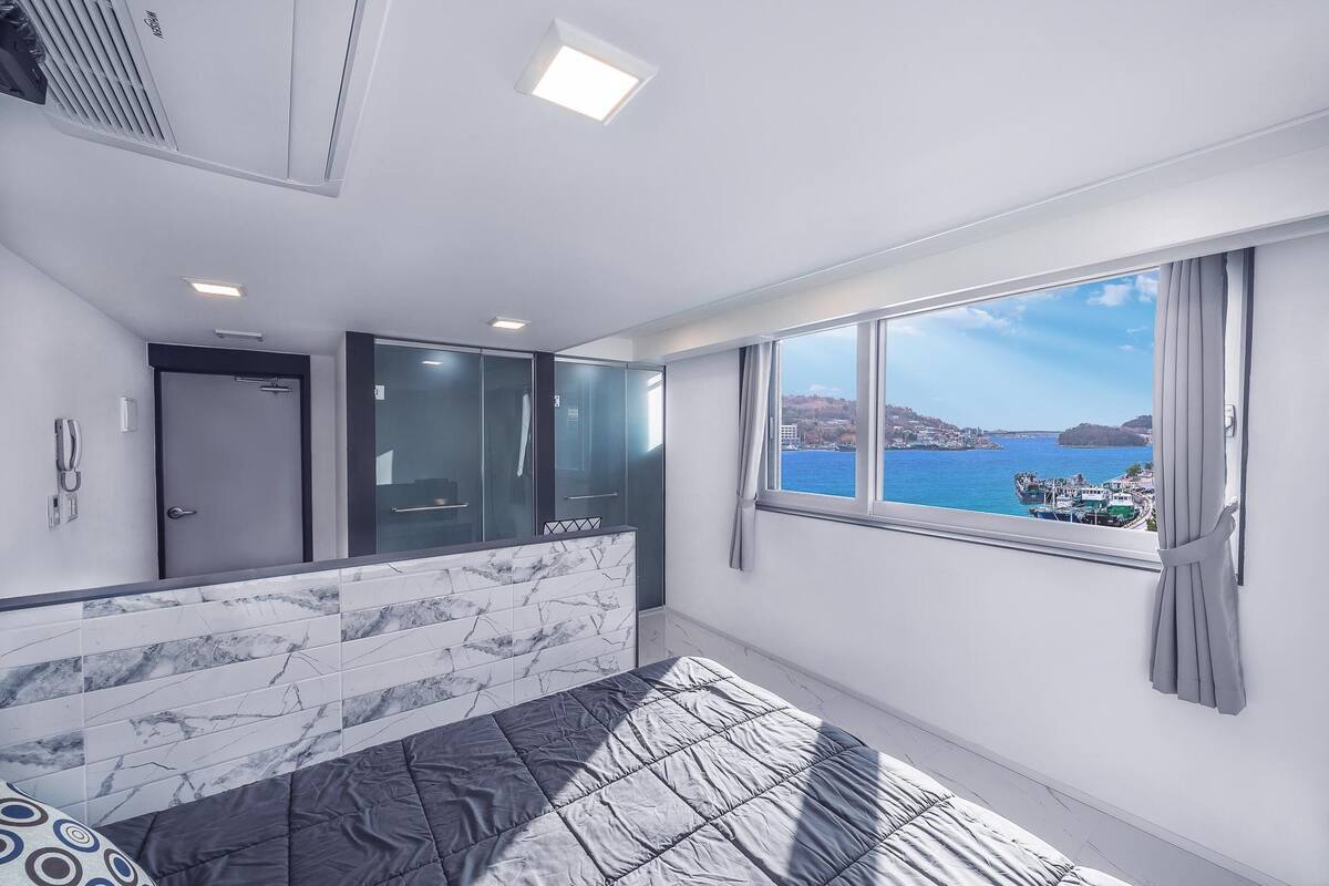 A modern bedroom with a large bed, a bathroom with glass doors, and a view of the sea.