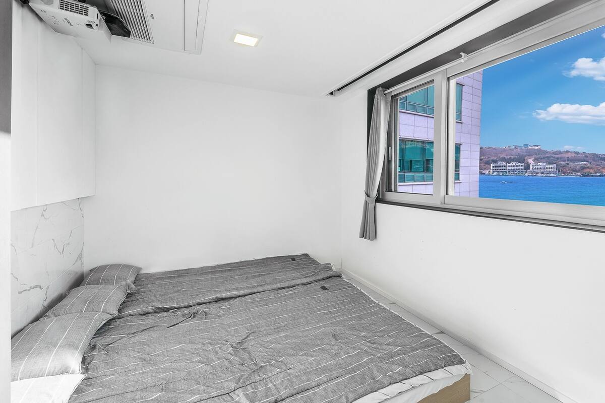 A small, unadorned room with a single bed, a window with a view of a distant coastline, and a ceiling vent.