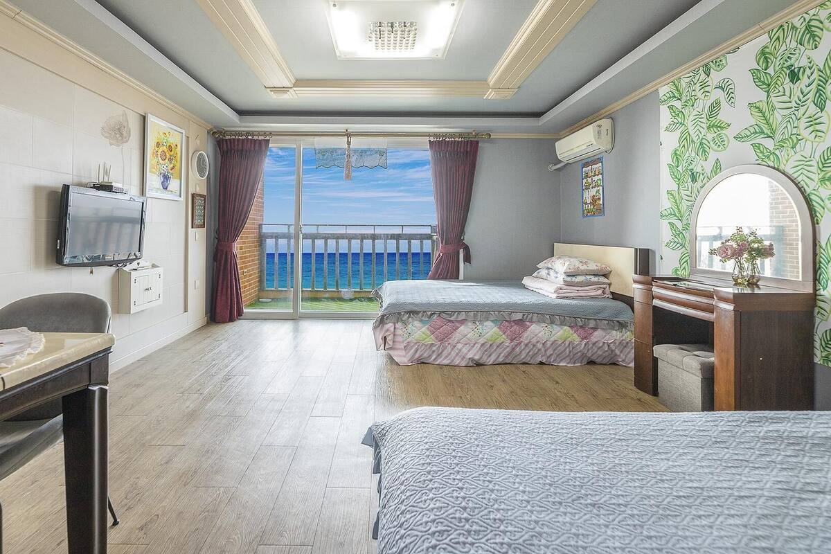 A hotel room with a bed, a desk, a TV, a dresser, and a balcony with a view of the sea.