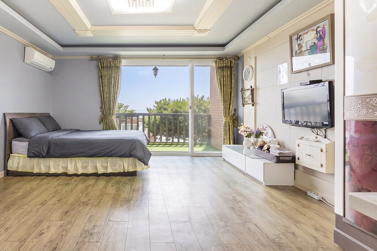 A bedroom with a bed, a TV, a balcony with curtains, and a wall-mounted clock.
