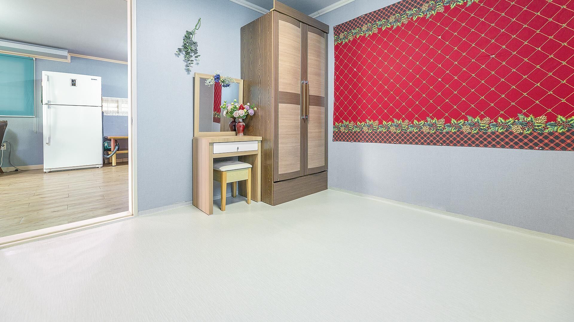 A room with a wooden wardrobe, a small desk with a chair, a white refrigerator, and a red patterned wall.