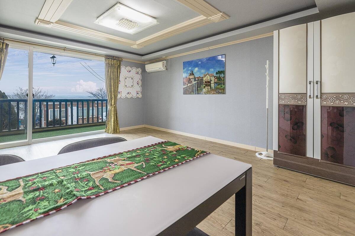 A room with a large window, a table with a green rug, a painting, and a wardrobe with floral designs.
