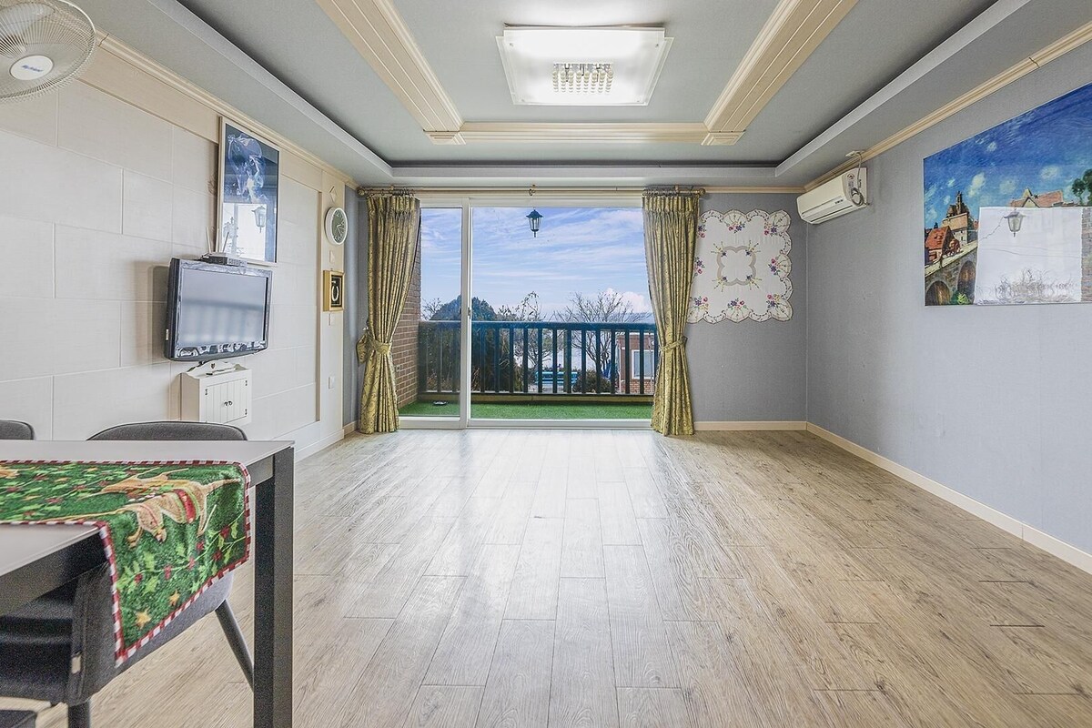 A room with a television, a table with a tablecloth, a balcony with a view, and a painting on the wall.