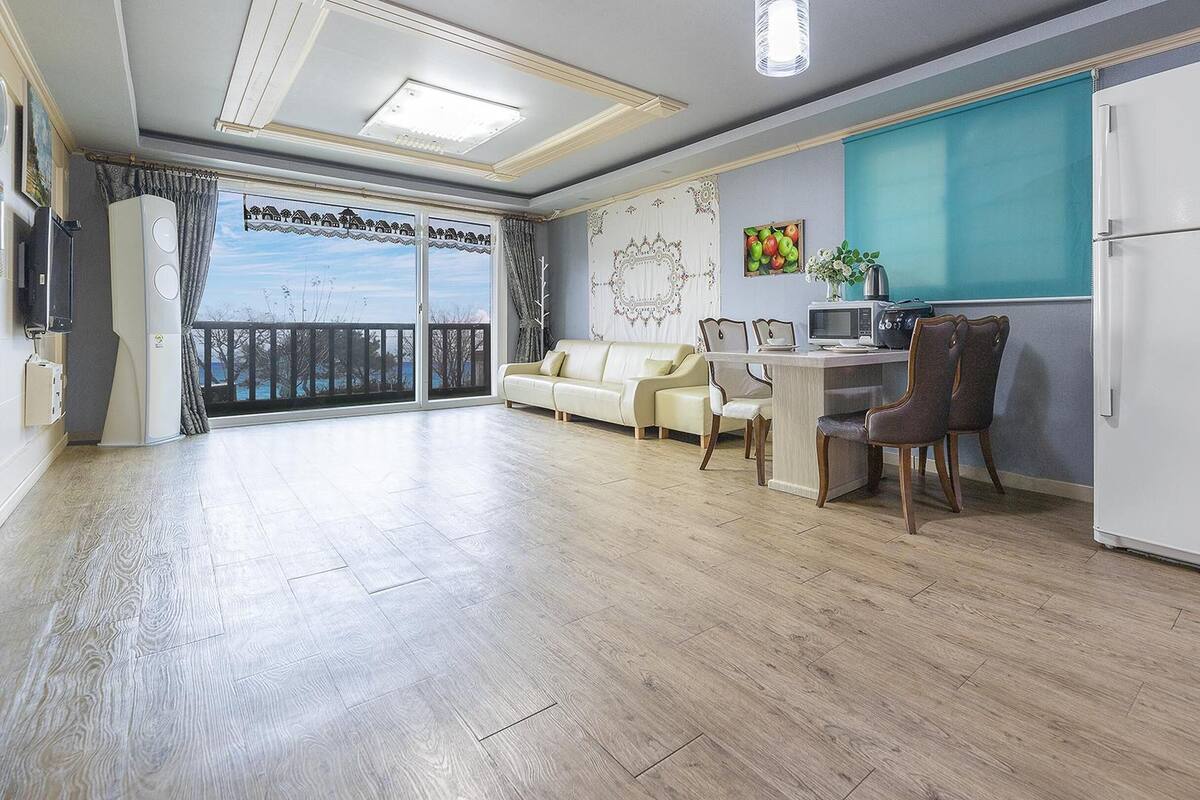 A spacious living area with a balcony, a dining table with chairs, a sofa, and a white refrigerator.