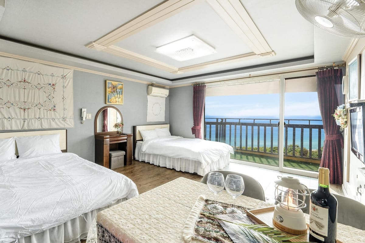 A hotel room with two beds, a large window with a view of the sea, a wooden desk, and a small table with glasses and a bottle.