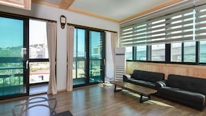 A spacious living room with a large sliding glass door leading to a balcony, a black leather sofa, a wooden coffee table, and a built-in air conditioning unit.