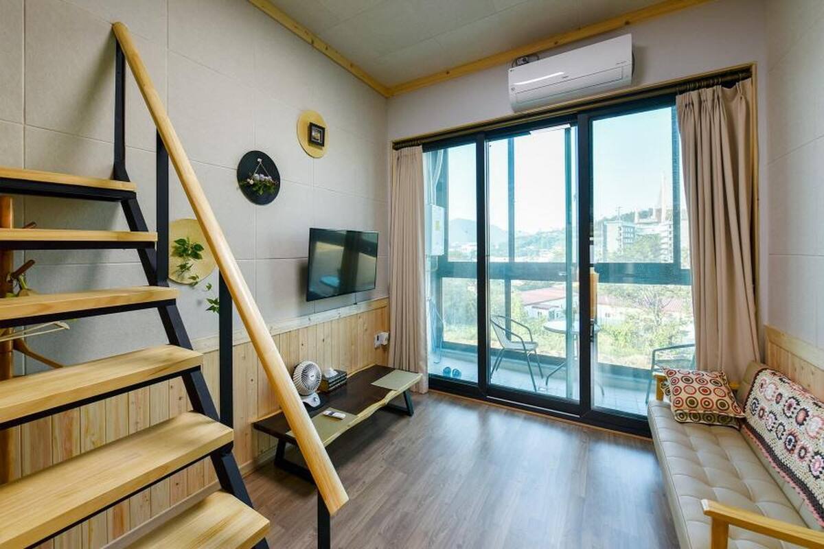 Una sala moderna con una escalera de madera, un televisor de pantalla plana, un sofá con cojines estampados y un amplio ventanal corredero que da a un balcón.