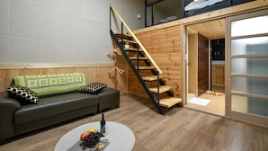 A compact living space with a sofa, a coffee table with fruit, a wooden staircase, and a sliding glass door leading to a bathroom.